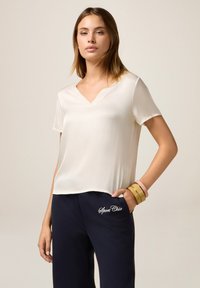 Camisa ligera de color crema con escote en v y mangas cortas, combinada con pantalones de chándal azul marino con bordado "Sport Chic". Textura suave.
