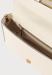 Beige bőrtáska sima felülettel, aranyszínű fémrészletekkel, cipzáras zsebbel és diszkrét márkázással a belsejében. Felső mágneses záródás.