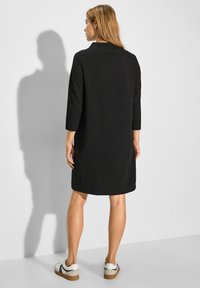 Schwarzes, strukturiertes Kleid mit Dreiviertelärmeln, lockerer Passform und rundem Ausschnitt. Tragen mit weißen Sneakers mit schwarzen Akzenten.