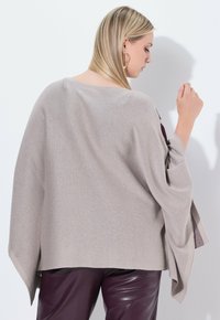 Poncho de punto gris claro, de gran tamaño, con mangas anchas, que presenta un ajuste holgado y una textura sutil. Combinado con pantalones de cuero ciruela brillantes.