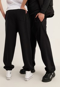 Black, loose-fitting joggers with elastic cuffs and side pockets, styled with white and black sneakers. Smooth cotton blend fabric.