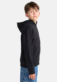 Felpa nera con zip e tasca a marsupio, realizzata in stoffa morbida. Presenta un grande cappuccio e polsini a coste, indossata con jeans in denim blu.