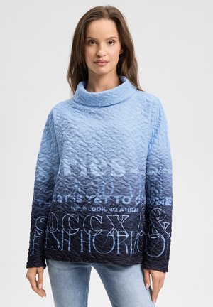 Frau trägt einen strukturierten, gesteppte Blau-Ombré-Rollkragenpullover mit gemischtem weißem und schwarzem Text, kombiniert mit hellblauen Jeans, vor weißem Hintergrund.