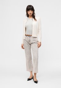 Nő áll krémszínű kardigánban, világosszürke felsővel, savmosott farmernadrágban, barna övvel és fekete, hegyes orrú lapos cipőben, fehér háttér előtt.