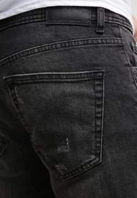 Detalle de un bolsillo trasero y la cintura de unos vaqueros negros desgastados, con una camisa blanca metida por dentro.