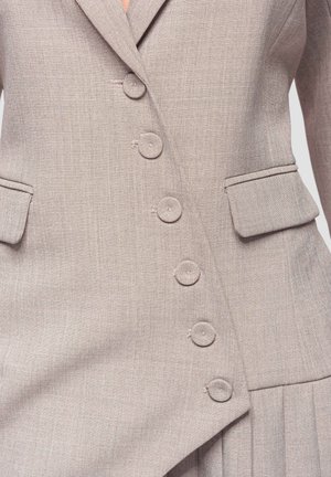 Blazer beige sartoriale con chiusura asimmetrica a bottoni e tasche con patta, che mostra un tessuto intrecciato texturizzato e una vestibilità strutturata.