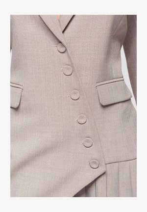 Blazer beige sartoriale con chiusura asimmetrica a bottoni e tasche con patta, che mostra un tessuto intrecciato texturizzato e una vestibilità strutturata.