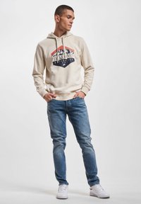 Cremefarbener Kapuzenpullover mit grafischem Logo, kombiniert mit hellblauen Jeans und weißen Sneakern. Geschmeidige Textur, lässige Passform.