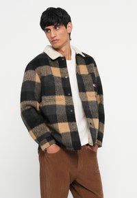 Black and tan checkered flannel shirt with a cream collar, featuring two front pockets and a button-up design. Worn over a white tee.