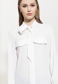 White blouse with a bow tie collar, two chest pockets, long sleeves, and a smooth, lightweight fabric texture.