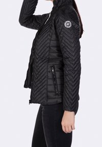 Doudoune noire avec motif en chevrons, poches zippées, col montant et patch logo sur la manche. Fini lisse et mat.