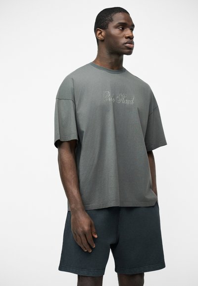 Homme portant un T-shirt gris surdimensionné avec le texte "Be Kind" et un short sombre, debout devant un fond clair uni, regardant sur le côté.