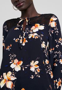 Blusa floreale con sfondo blu navy, caratterizzata da fiori arancioni e bianchi. Dettagli in pizzo sulle spalle e chiusura con lacci al collo.
