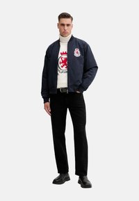 Chaqueta bomber de la Marina con emblema bordado, superpuesta a un suéter de cuello alto blanco con un logo de león rojo. Pantalones negros y botas.