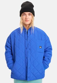 Modrá prošívaná bunda se vzorem rombů, s předním zapínáním na knoflíky a výraznou náprsní kapsou s logem. Na vrchu černá pletená čepice.