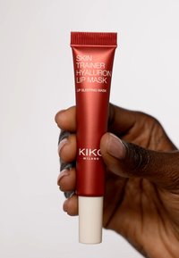 Rød tube med hvit topp, merket "Skin Trainer Hyaluron Lip Mask." Hånd som holder tuben, som fremhever det stilige designet og den blanke overflaten.