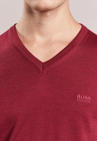 Burgundy V-ringad tröja gjord av ribbat tyg, med en liten broderad "BOSS"-logotyp i det nedre vänstra hörnet.