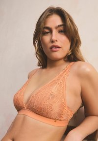 Bralette in pizzo di un leggero arancione con coppe triangolari, motivo floreale intricato, ampia fascia elastica e sottili spalline regolabili.