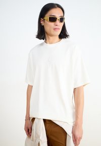 Witte T-shirt met korte mouwen en een subtiele logo, gecombineerd met een bruine broek. Het model draagt gele zonnebril met een gestructureerd frame.