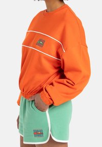 Felpa arancione con polsini a coste e strisce bianche, abbinata a pantaloni corti verde menta con logo e bordo bianco.