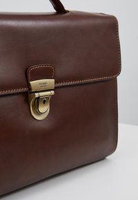 Bolso de cuero marrón con un diseño estructurado, que presenta un cierre de latón y costuras detalladas a lo largo de los bordes. Textura suave, hardware mínimo.