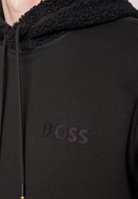 Gros plan d'un sweat à capuche noir avec capuche texturée et logo "BOSS" embossé sur la poitrine, avec des cordons aux extrémités noires et orange.