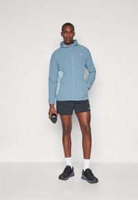 Helle blaue Kapuzenjacke mit einem Frontreißverschluss, kombiniert mit schwarzen Shorts und Sportschuhen. Hält eine Wasserflasche und steht vor einem neutralen Hintergrund.