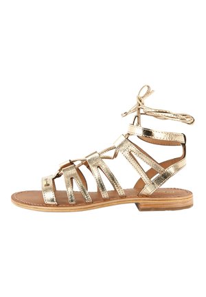 Złote metaliczne płaskie sandały z wieloma paskami i wiązaniem w okolicy kostki. Teksturowane wykończenie, płaska podeszwa i brązowa wkładka.
