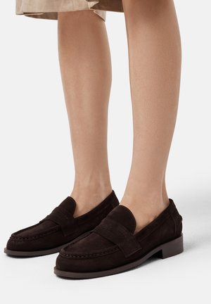 Braune Wildlederslipper mit runder Zehenpartie, ausgestattet mit einer genähten Detail und einem kleinen Blockabsatz. Das Design umfasst einen auffälligen Riemen über der Vorderseite.