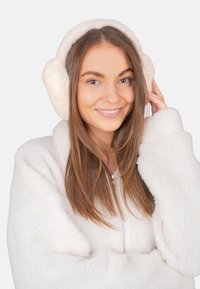 Cache-oreilles en fausse fourrure blanche sur une personne aux longs cheveux bruns, portant une veste blanche en peluche à fermeture éclair, souriant et touchant les cache-oreilles.