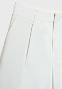 Pantalon blanc sur mesure montrant la taille avec passants de ceinture, plis avant et poches latérales sur un fond clair.