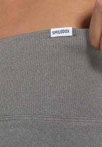 Grijze geribde stof met een rekbare textuur, met een SMILODOX-label op de bovenrand genaaid; close-up van de details van de tailleband.