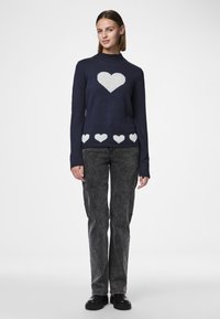 Marineblå strikkegenser med et stort hvitt hjerte mønster på brystet og mindre hjerter langs kanten. Lange ermer og ribbestrikket hals.