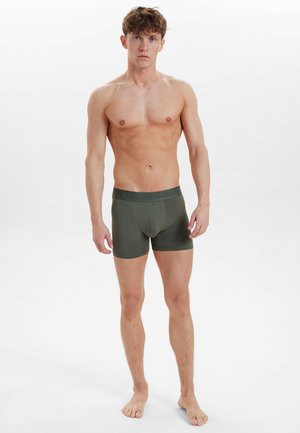 Grønne bomuldsboxershorts med et tætsiddende design, elastisk talje og en glat tekstur, båret af en model stående barfodet.