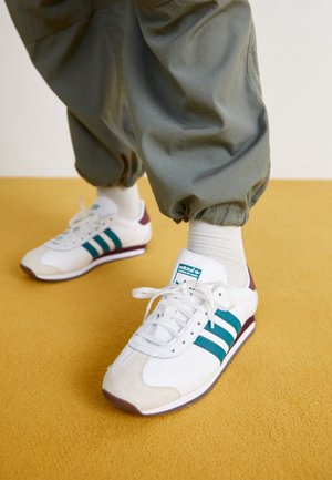 Białe sneakersy Adidas z turkusowymi paskami i bordowymi detalami, noszone z białymi skarpetkami i luźnymi zielonymi spodniami na żółtej podłodze.
