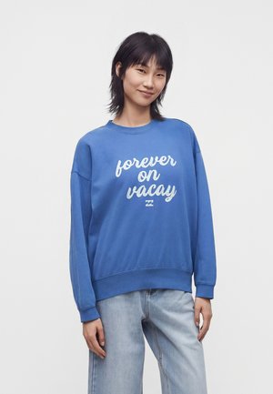 Persoană tânără cu păr închis la culoare, purtând un hanorac albastru cu textul „forever on vacay” și blugi bleu deschis pe un fundal alb simplu.