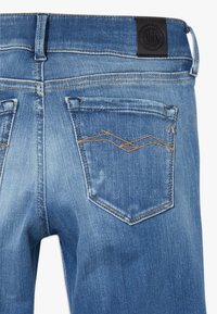 Vaqueros de mezclilla azul con un acabado desgastado, presentan costuras marrones, dos bolsillos traseros y una etiqueta de cuero negra en la cintura.