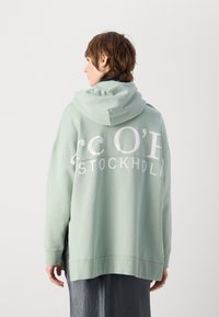 Overmaatse mintgroene hoodie van zacht materiaal, met op de achterkant een grote witte opdruk van "c O' H STOCKHOLM" en een capuchon met trekkoord.