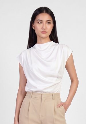 Femme aux longs cheveux foncés portant un top blanc drapé sans manches et un pantalon beige taille haute, posant avec une main dans la poche devant un fond uni.