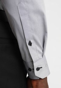 Vaaleanharmaa kauluspaidanshiis, jossa mustat napit ja teksturoitu kangas. Kalvosimet ovat jämäkät, kontrastivärisillä tummilla yksityiskohdilla.