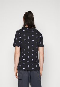 Henkilö, jolla on tummat hiukset poninhännällä, pukeutuneena mustaan t-paitaan, jossa on violetteja Nike-logoja ja "Just Do It" -tekstikuvioita, yhdistettynä tummaan alaosaan.