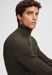 Dunkelgrüner gerippter Rollkragenpullover mit hohem Kragen. Figurnaher Schnitt, strukturierter Stoff und dezente Nahtdetails an den Schultern.