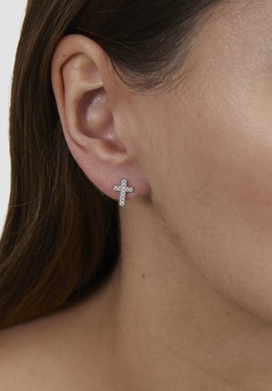 Close-up of a person’s ear wearing a small cross-shaped earring studded with clear stones.