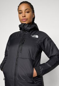 Žena nosi crnu izoliranu jaknu North Face s zipovima na džepovima i visokim okovratnikom, stoji naslonjena na ravnu svijetlu pozadinu.