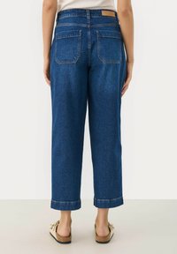 Blå denimbukser med vida ben och uppvikta fållar, två bakfickor och en tanfärgad läderlapp vid midjan. Slät textur, avslappnad passform.