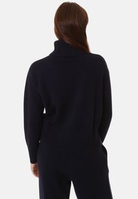 Donna con lunga chioma castana, che indossa un maglione a collo alto blu scuro e pantaloni abbinati, vista di spalle contro uno sfondo semplice.