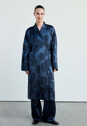 Femme portant une longue robe en satin bleu foncé avec des motifs floraux noirs par-dessus un pantalon large assorti et des chaussures noires.