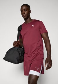 Diepgrijze sportshirt en shorts met witte logo; model draagt een zwarte sporttas. Shorts hebben een witte accent en zijsplitten. Glad stoftextuur.
