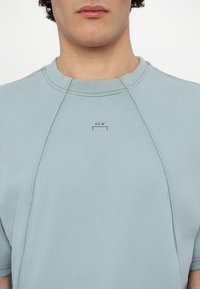 Sweat-shirt bleu clair à manches courtes avec un col rond, présentant des coutures contrastées et un petit logo imprimé sur la poitrine.