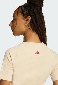 Beżowy bawełniany t-shirt z raglanowymi rękawami, z różowym logo Adidas na karku. Gładka faktura, luźny krój.